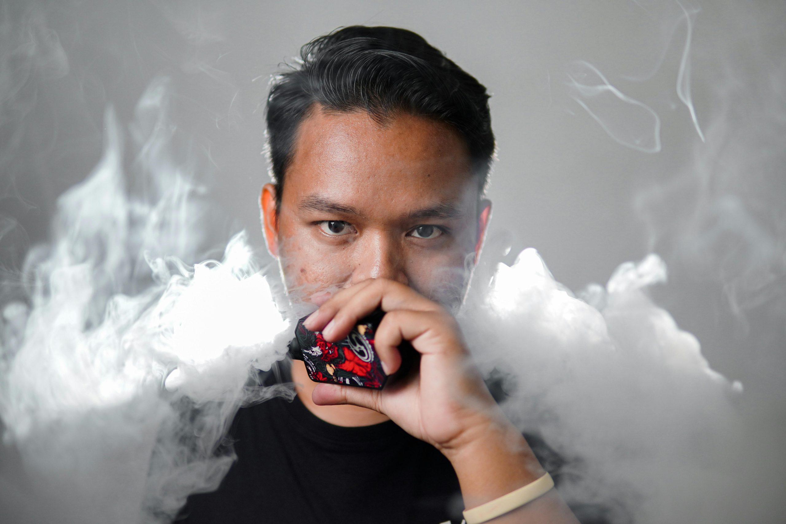 Portrait of a young man with black hair vaping, surrounded by smoke against a gray background.