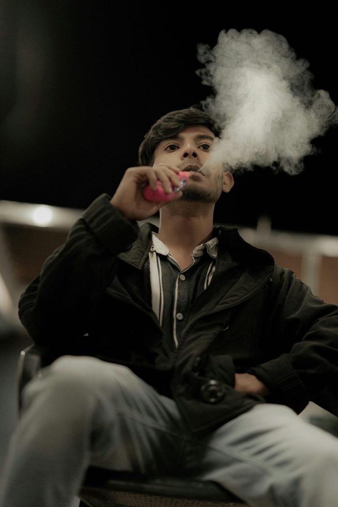 A young man exhaling vapor from an e-cigarette, set against a moody indoor backdrop at night.
