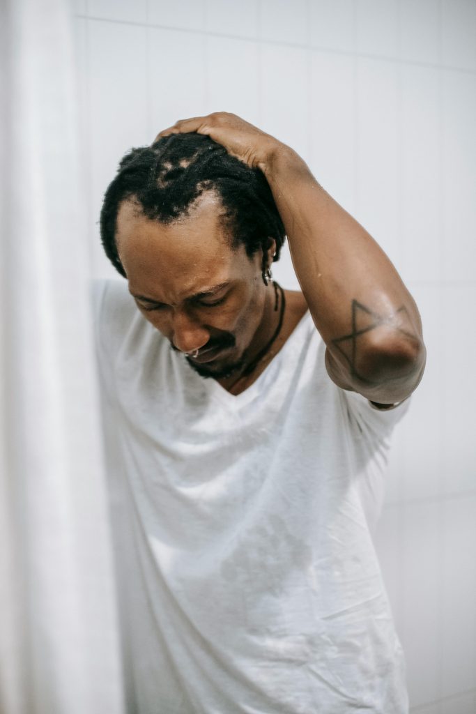Preoccupied frustrated African American male in white shirt touching head nervously in shower and looking down in unpleasant thoughts