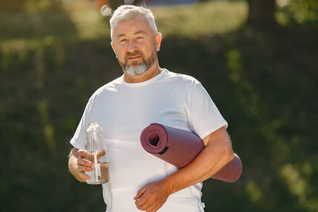 Senior man holding a yoga mat and water bottle outdoors, ready for exercise.
