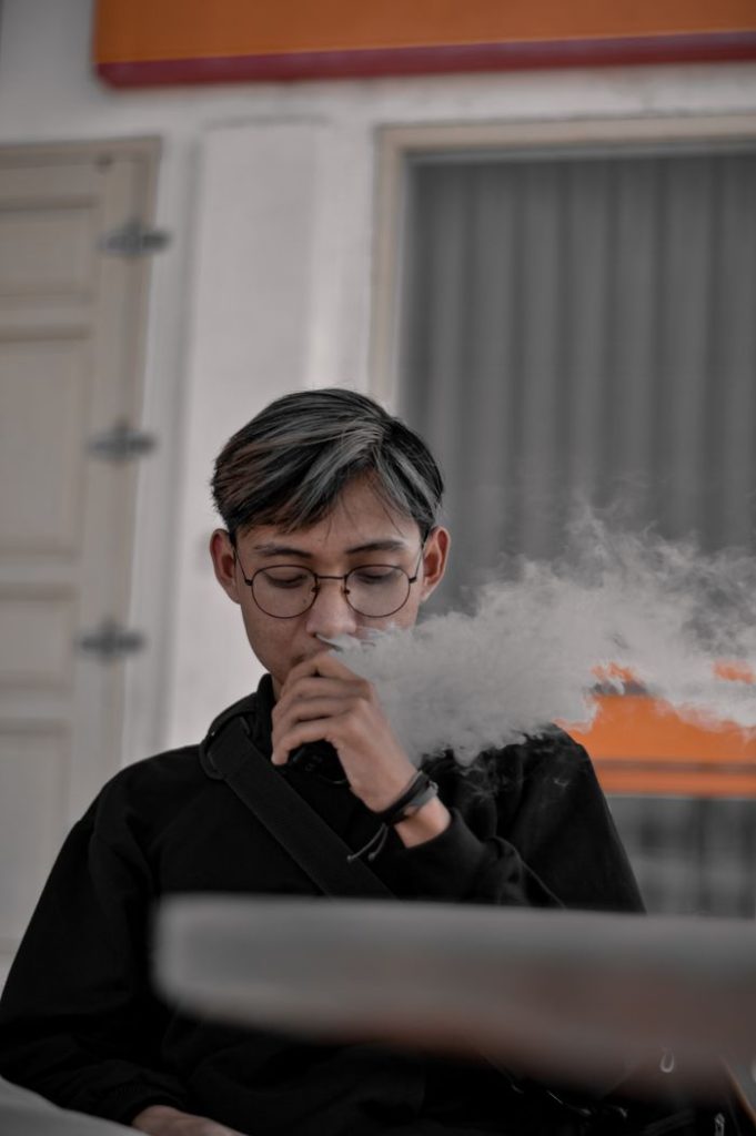 Portrait of a young adult wearing glasses while vaping indoors, producing a visible cloud of vapor.