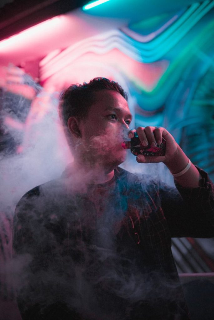 Asian man vaping in a colorful and smoky nightclub with vibrant neon lights.